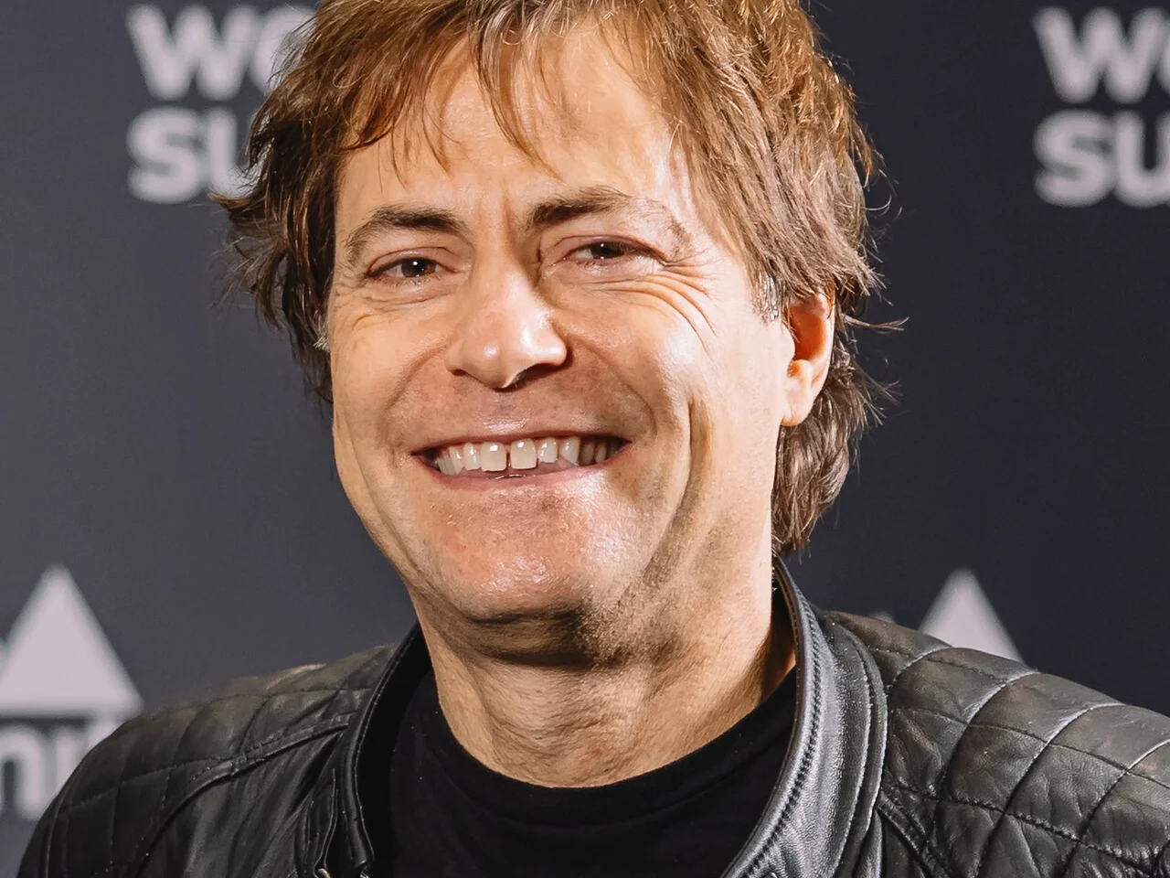 Max Tegmark i svart skinnjacka ler framför en fotovägg på Web Summit