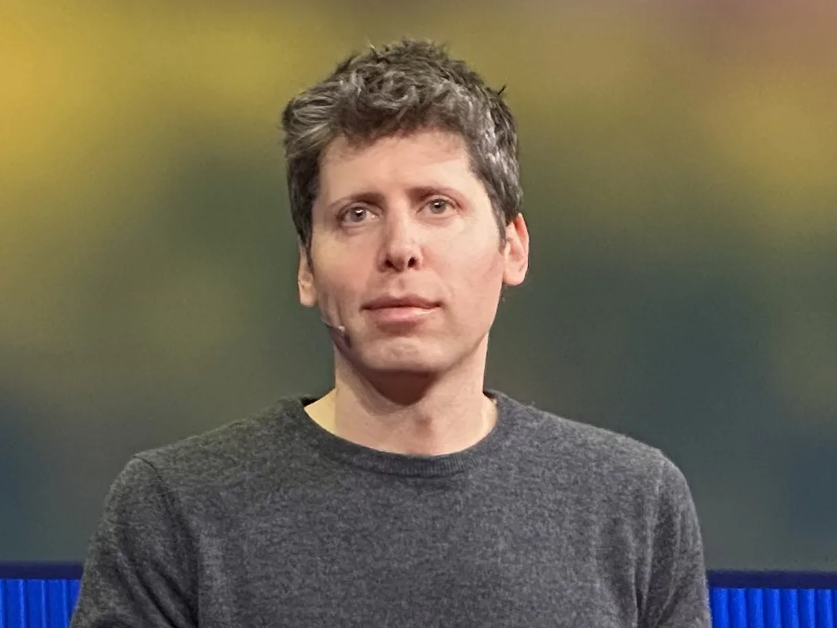 Sam Altman i grå tröja med headsetmikrofon på scen under TED